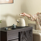 The cream SMEG Retro Stovetop Kettle sitting on a dark wood tea cabinet, ready for pouring, with a person seated on a sofa in the background creating a warm, at-home ambiance.